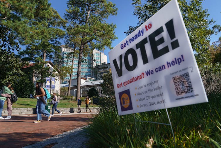 A large white sign says 'Vote!' People walk past the sign outside, in what appears to be a green campus with trees.