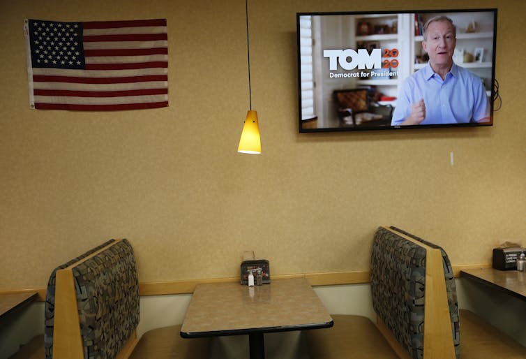 A TV on a wall above a restaurant booth shows a white man in a blue shirt with the words 'Tom 2020 Democrat for president' next to him.