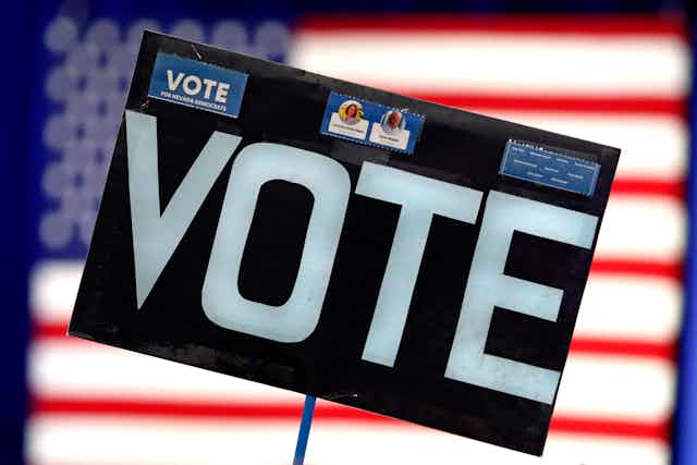 A large black sign says vote in white font, with an American flag blurred in the background behind it.