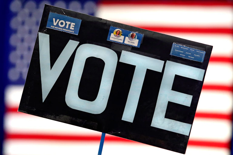 A large black sign says vote in white font, with an American flag blurred in the background behind it.