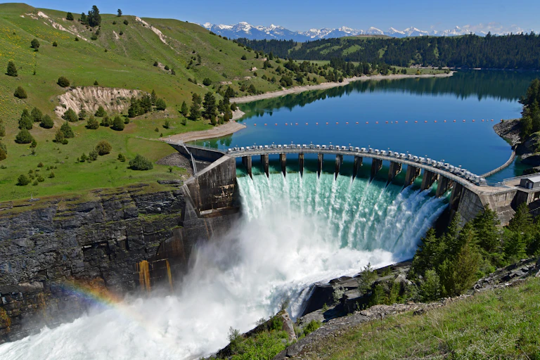 Water from a lake spills over a dam in the foreground with a pine forest in the background and snow-covered mountains in the distance