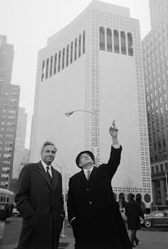 Two white men in suits, ties and coats stand next to each other in New York in front of a tall building. One of them is pointing and looking up