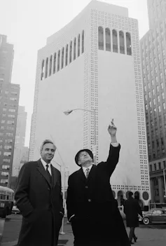 Two white men in suits, ties and coats stand next to each other in New York in front of a tall building. One of them is pointing and looking up