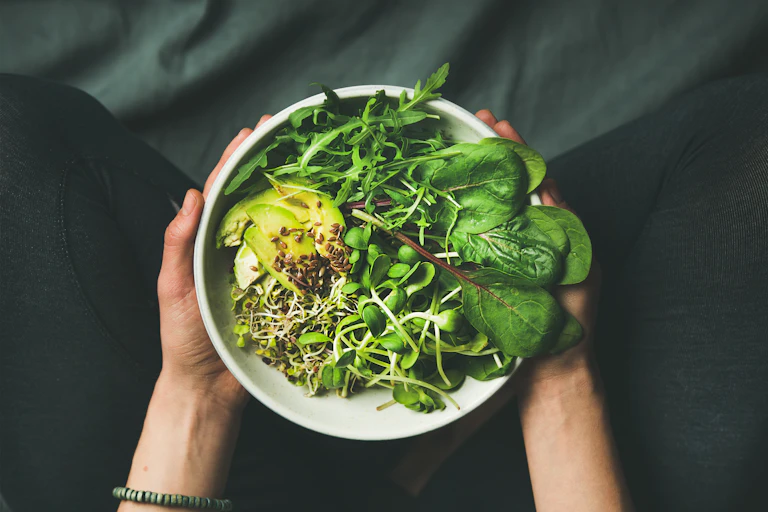 A woman holds a plate of raw vegan foods, including spinach, arugula, nuts, and avocado.