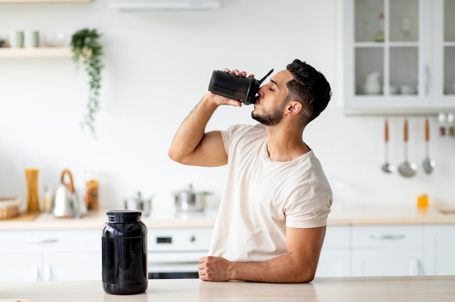 A fit young man drinks a protein shake in his kitchen.