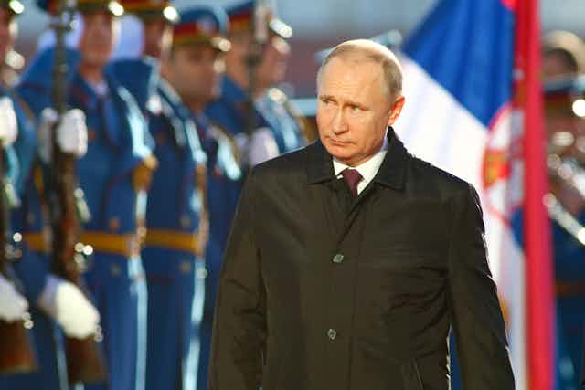 Soliders in a line standing at attention in front of a Serbian flag and Vladimir Putin.