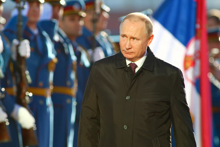 Soliders in a line standing at attention in front of a Serbian flag and Vladimir Putin.