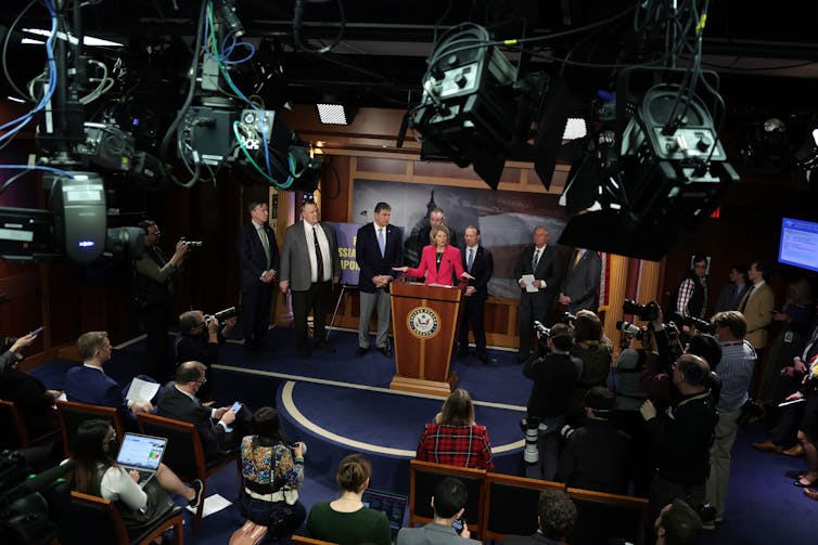 A woman in a red jacket speaks from a lectern, surrounded by half a dozen others and being filmed by large cameras.