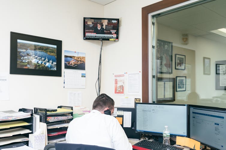 A man in a white shirt speaks into a telephone in front of a bank of computer screens.