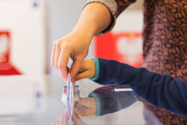 A woman's hand and a small child's hand put a vote into a ballot box together