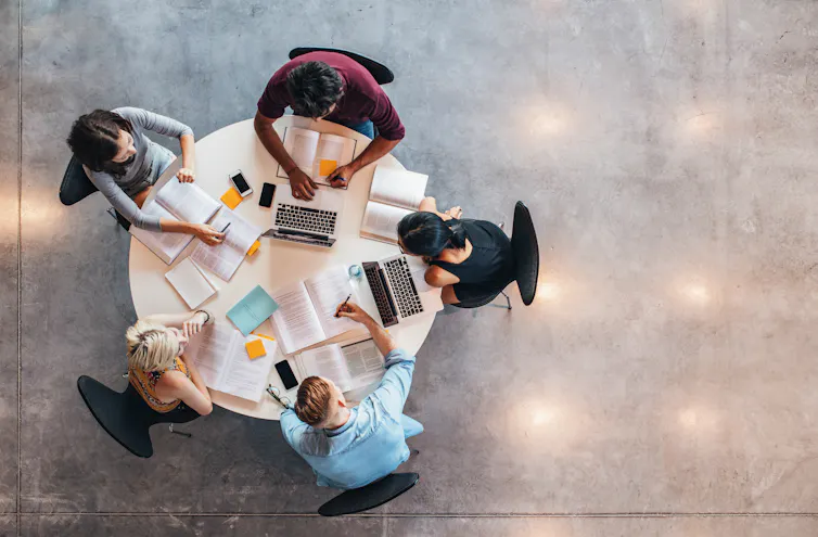 A bird's eye view of four people sitting at a round table with laptops and notebooks.
