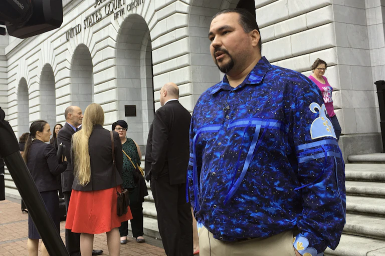 A man in a blue patterned shirt stands in front of a large building.