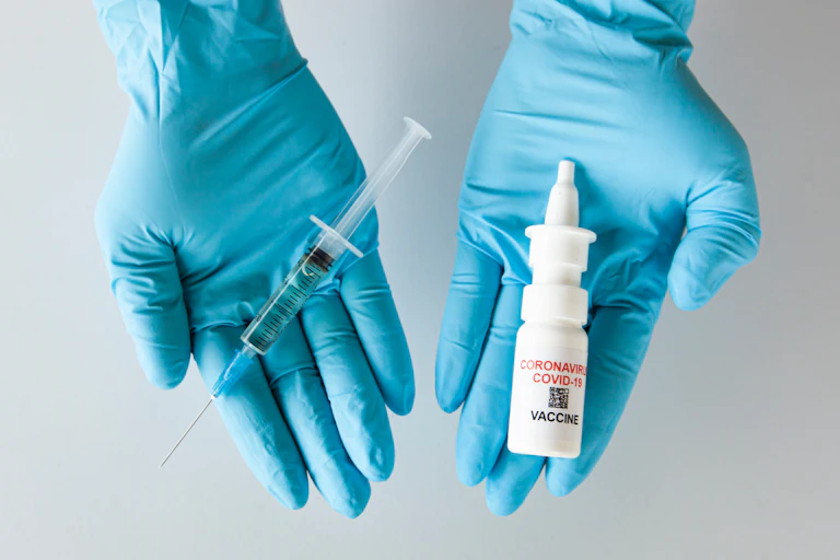 Two gloved hands: one holds a traditional vaccine, and the other a nasal spray.