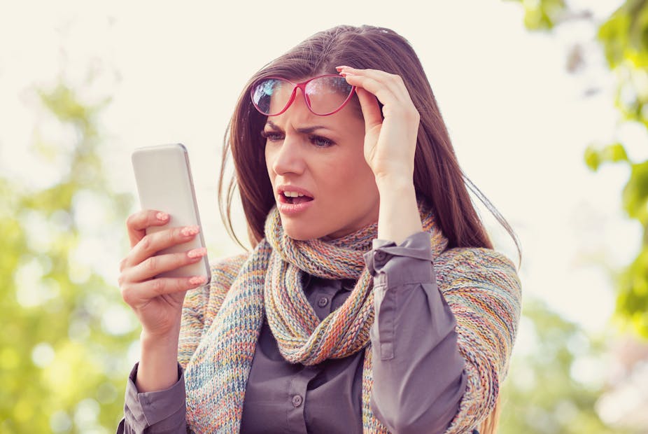 A woman lifts up her glasses and looks in shock and confusion at her mobile phone