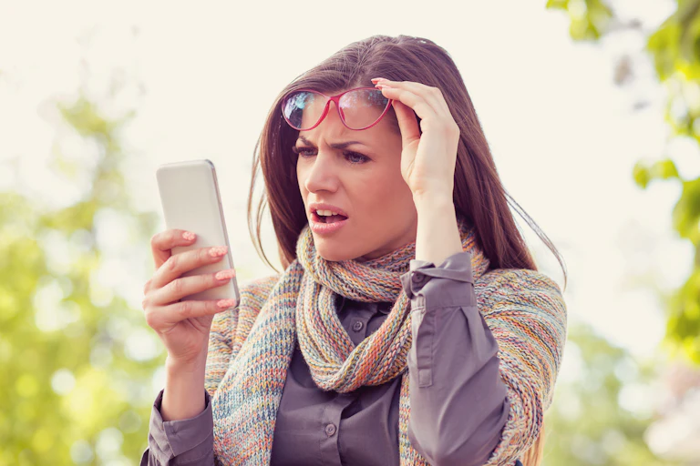 A woman lifts up her glasses and looks in shock and confusion at her mobile phone