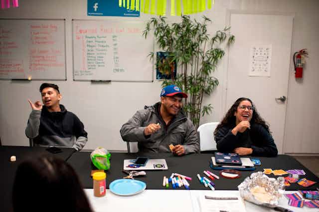 Three people laugh as they sit at a table in a conference room, with markers and snacks scattered around them.