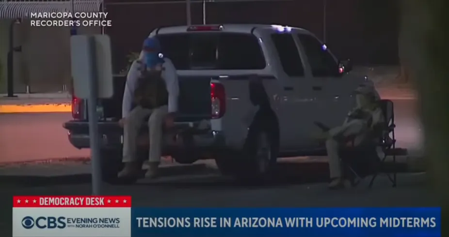 People with hats, masks and tactical vests sit near a pickup truck in a parking lot.