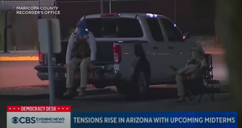 People with hats, masks and tactical vests sit near a pickup truck in a parking lot.