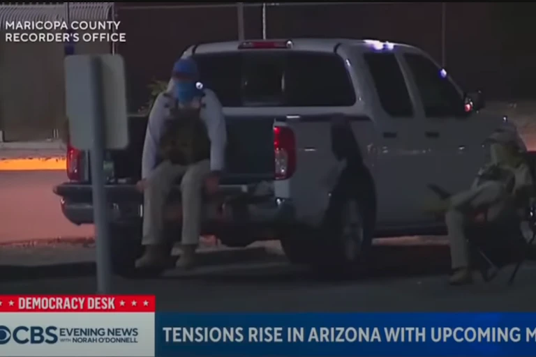 People with hats, masks and tactical vests sit near a pickup truck in a parking lot.