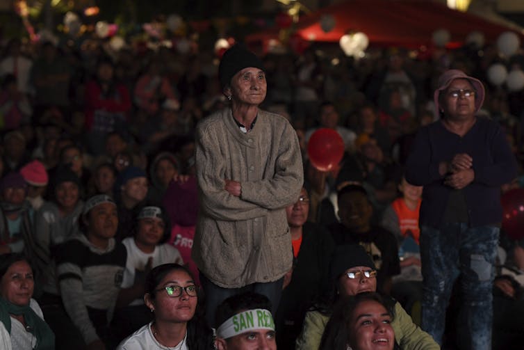 A few people stand up amid a tightly packed crowd of people sitting in the dark.