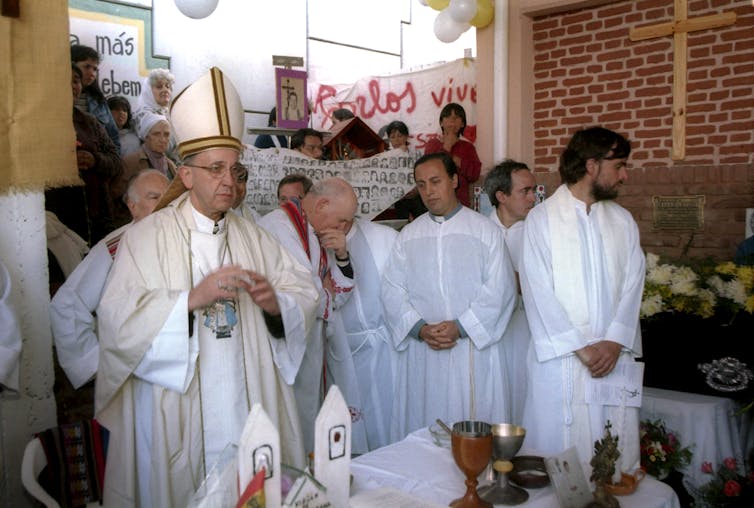 Catholic priests, one of them wearing a tall hat, stand in white robes in front of a brick wall with a wooden cross on it.