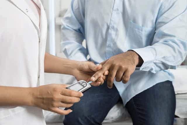 Cropped image of hands of a health-care worker in white coat using a glucometer on the finger of a person in a blue shirt.
