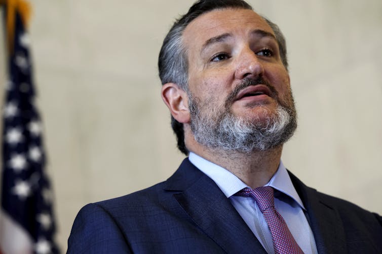 A man with a graying beard dressed in a business suit is seen talking with the American flag in the background.