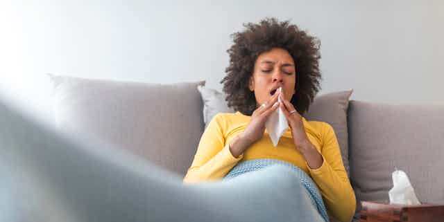 A person is sitting on a couch with a blanket over their legs. They are mid-sneeze.