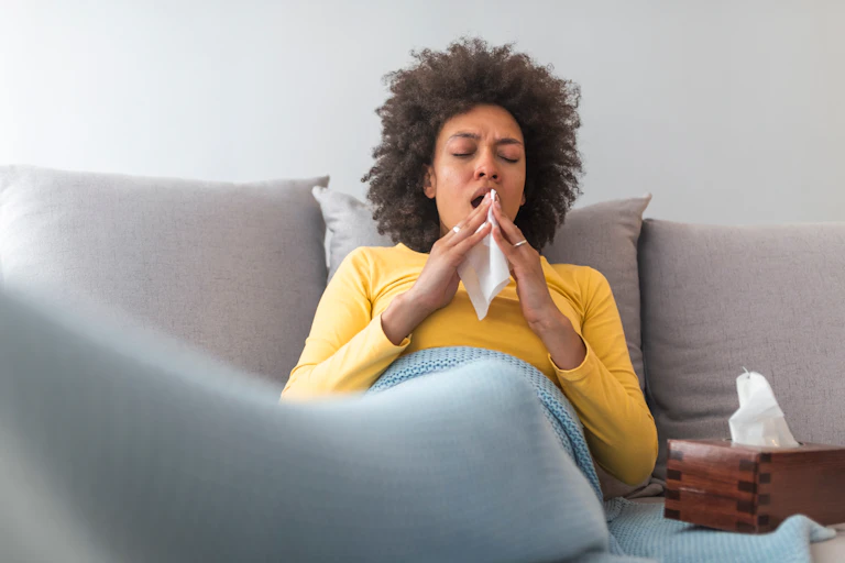 A person is sitting on a couch with a blanket over their legs. They are mid-sneeze.