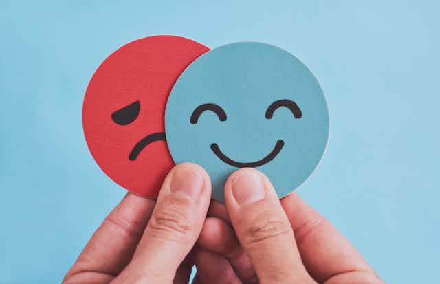 Hands holding two paper cutouts with a blue smiley face partially hiding a red sad face