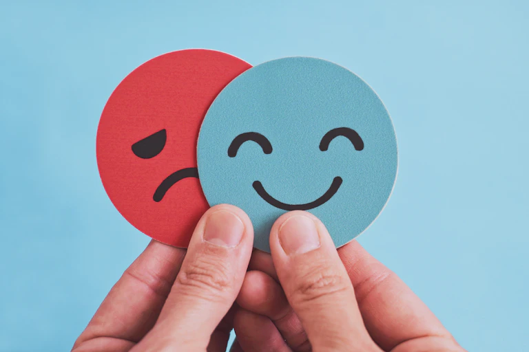 Hands holding two paper cutouts with a blue smiley face partially hiding a red sad face