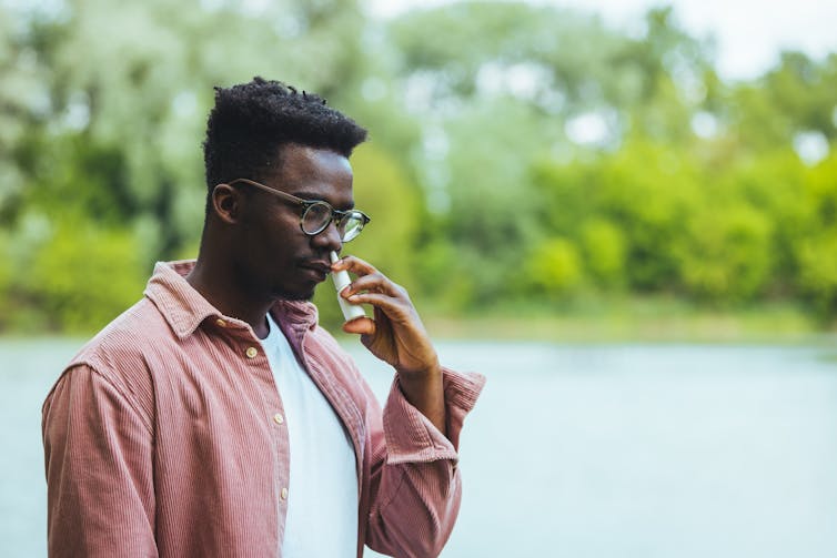 A person is standing outside using nasal spray. They wear glasses.