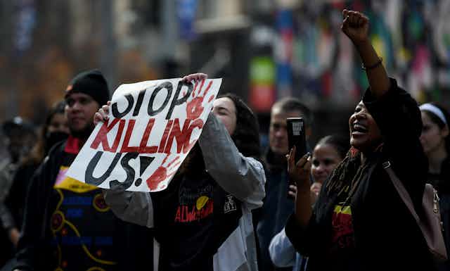 Indigenous protestors hold a sign: 'STOP KILLING US!'.