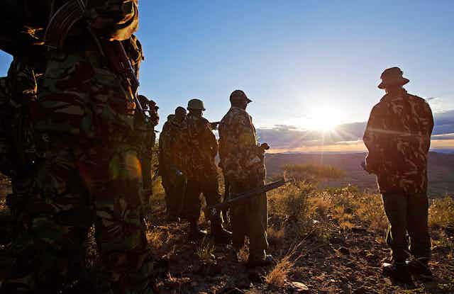 Men in police uniform and holding guns look out into a valley as the sun sets in a wilderness.