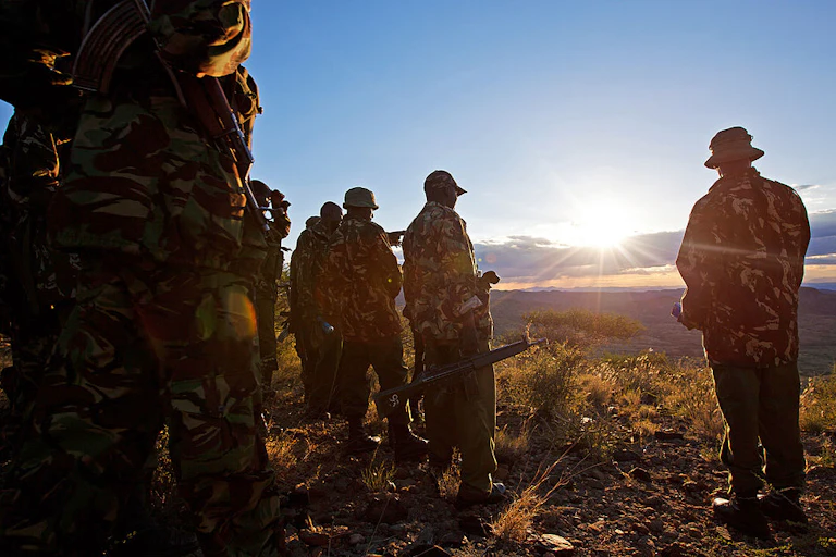 Men in police uniform and holding guns look out into a valley as the sun sets in a wilderness.