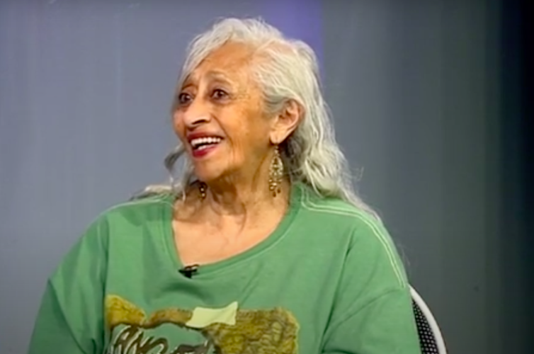 An elderly woman with long gray hair smiles as she speaks.