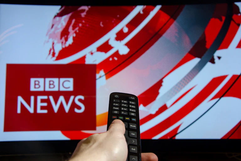 Hand points remote control at television showing BBC News broadcast