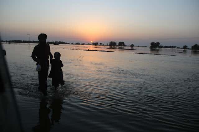 A parent and child wade through flood water at dusk.