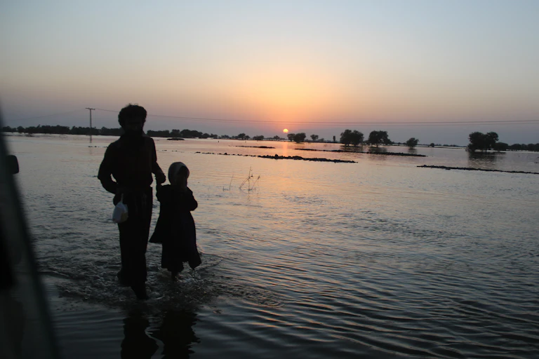 A parent and child wade through flood water at dusk.