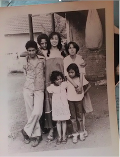 A black and white photo of a woman with pigtails surrounded by five children of varying ages. She has her hands around them and is smiling.