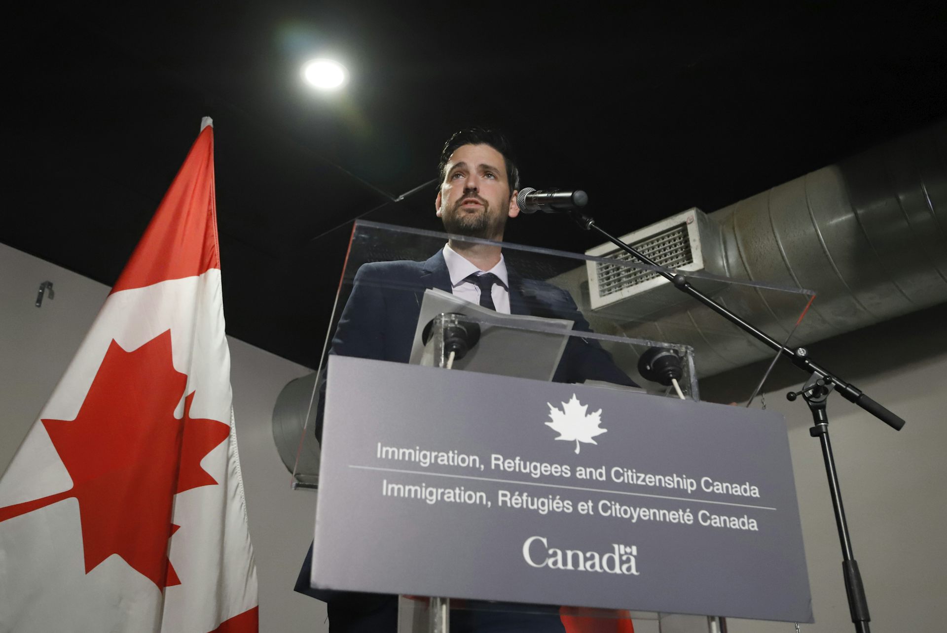 A man in a suit at a podium with a Canadian flag behind him.