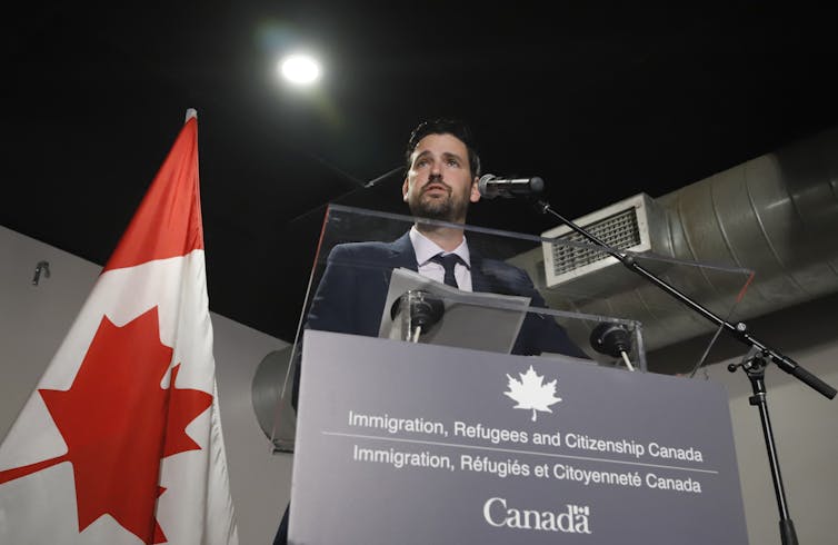 A man in a suit at a podium with a Canadian flag behind him.