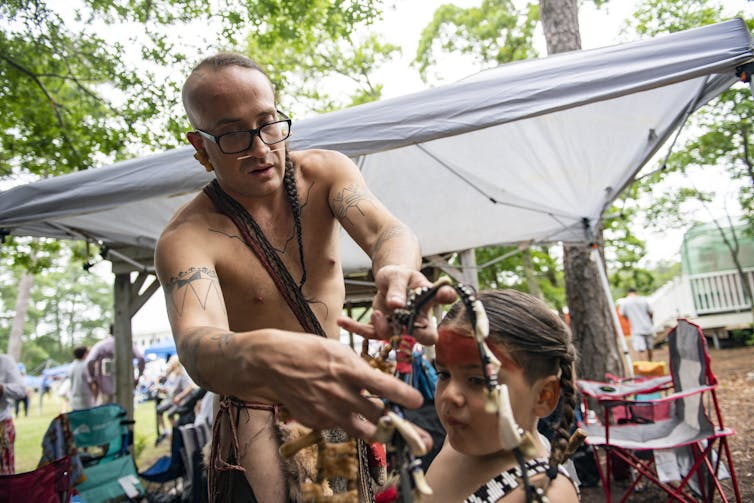 A man wearing a loincloth and glasses places a necklace over a child's head.