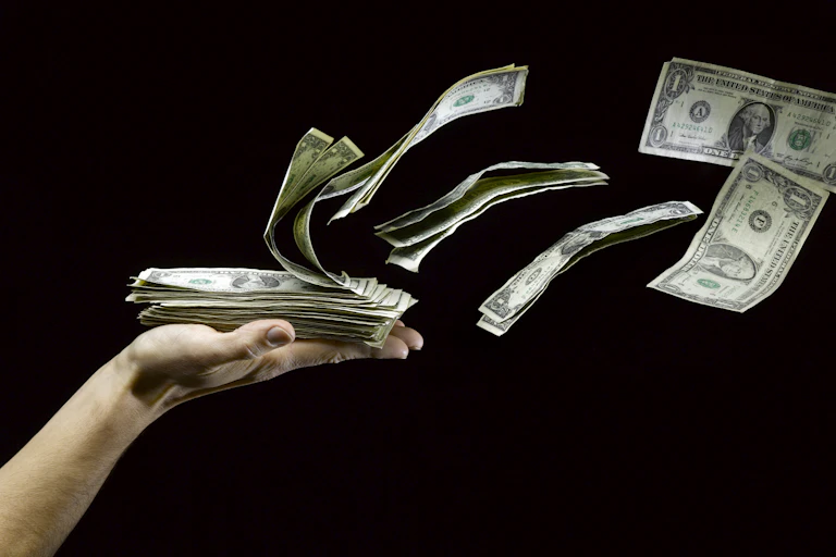 an outreached hand holds one dollar bills that are floating away as if being blown by the wind