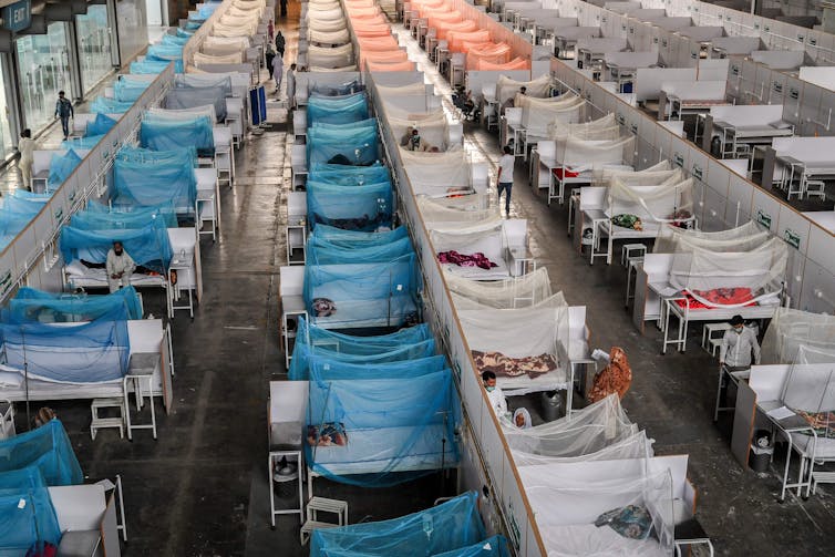 Rows of beds, some covered with mosquito nets, fill a warehouse-like space. Doctors visit with some of the patients.