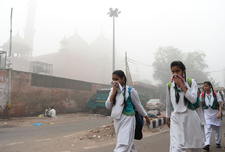 Three school girls with backpacks walk through smog along a road while covering their mouths with handkerchiefs.