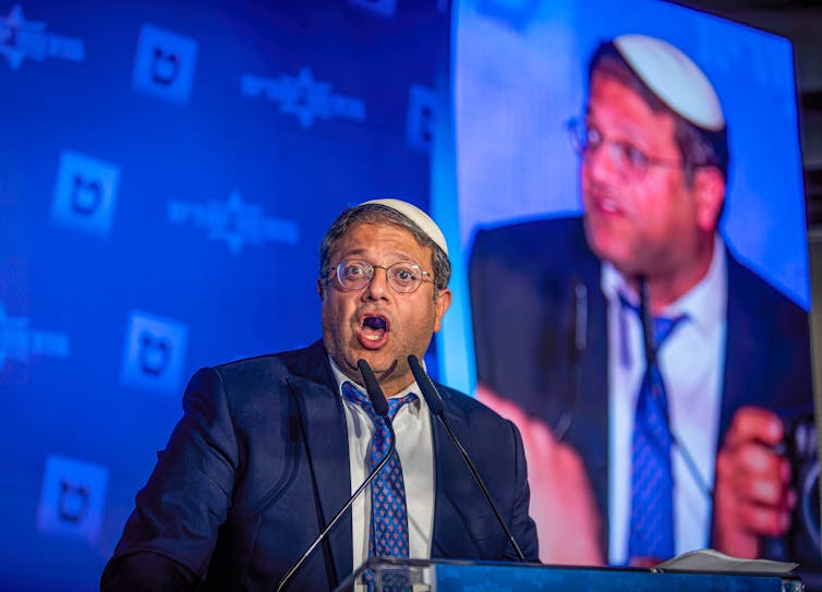 A man in glasses, suit and a yarmulke speaks into a microphone.