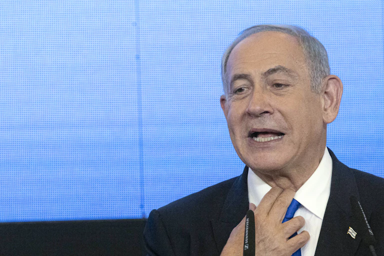 A man in a dark suit and blue tie grabs at his shirt collar.