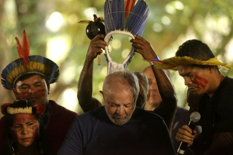 Indigenous men place a headdress on a grey-haired man whose head is bowed.