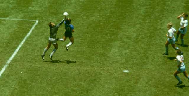 A soccer star in a blue and black kit outjumps a goalkeeper with his arm outstretched. England players in white look on.